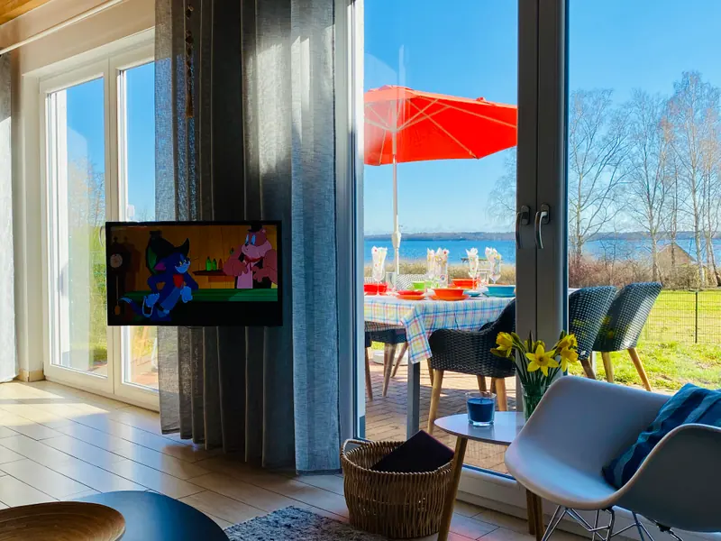 Blick vom Sofa durch die bodentiefen Fenster auf die Terrasse und den See